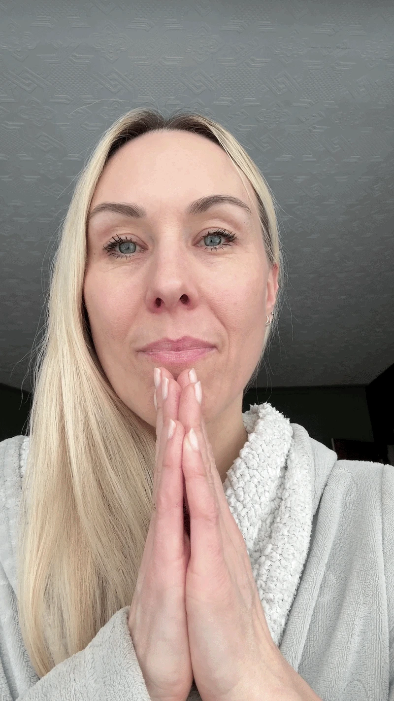 A person in a light robe looks towards the camera with their hands together